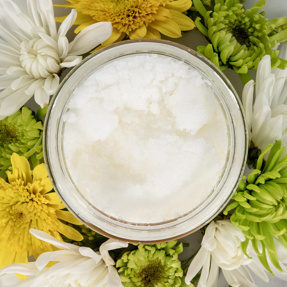 Birds eye view of an open jar filled with salt/oil combination. Jar is surrounded by green, yellow and white flowers